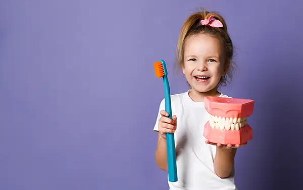 A smiling young girl holding up an oversized dental mouth model and toothbrush, showing excitement for kids dentistry.
